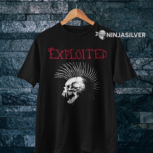 Pode incluir: T-shirt preto com um desenho de caveira e o nome da banda "The Exploited" em letras vermelhas. O design punk rock está impresso na t-shirt, pendurada num cabide de madeira contra uma parede texturizada.