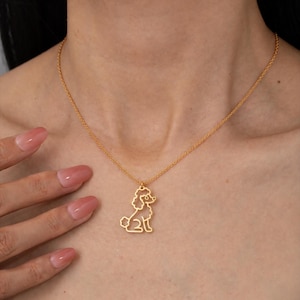 Könnte beinhalten: Eine goldfarbene Halskette mit einem zarten, offenen Drahtanhänger in Form eines sitzenden Pudels. Der Anhänger hängt an einer dünnen, passenden goldfarbenen Kette. Das Design zeichnet das lockige Fell, die Ohren und den Schwanz des Pudels nach.