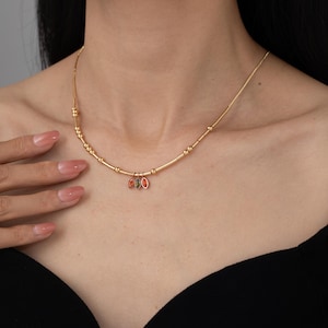 May include: A gold-toned necklace featuring a delicate chain with small cylindrical beads and three small marquise-shaped charms in orange, green, and red hues hanging from the center of the chain, resting against the collarbone area.