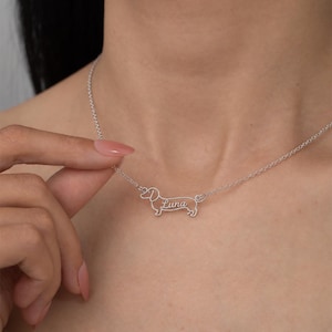 Könnte beinhalten: Eine silberfarbene Halskette mit einem Anhänger in Form eines Dackels. Die Silhouette des Hundes ist metallisch umrandet und enthält den Namen Luna in Schreibschrift in der Mitte des Körpers, befestigt an einer feinen Gliederkette.