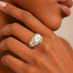 May include: A silver-toned ring featuring a smooth, irregular, organic-shaped white pearl set into a polished metal band. The ring has a wide, solid shank that tapers slightly towards the back, showcasing a high-shine finish and a sculptural design.