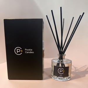 May include: A black box and a clear glass diffuser bottle with black reeds. The box and bottle have a white logo with the text "Pookie Candles". The bottle has a label that says "Flower Bomb".