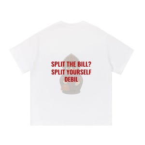 Puede incluir: Una camiseta blanca de manga corta con un gráfico de una muñeca rusa tradicional en tonos marrones y rojos. El texto impreso en letras mayúsculas rojas dice: SPLIT THE BILL? SPLIT YOURSELF DEBIL. El diseño está centrado en la camiseta.