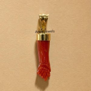 Puede incluir: Un colgante con una mano de coral rojo tallada suspendida de una anilla de metal dorado pulido. La pieza incluye el texto Anna Jewels impreso en negro cerca de la parte superior. El diseño muestra una orientación vertical con un puño detallado.