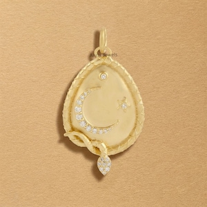 May include: A gold pendant shaped like a teardrop with a textured border. The centre features a crescent moon and a small star, both encrusted with clear stones. A small stone is set above the moon, and a snake-like tail wraps around the bottom.