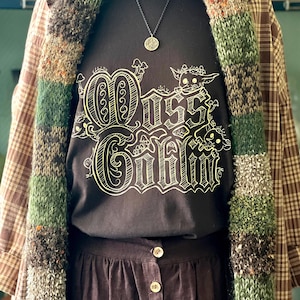 May include: A dark brown t-shirt with the text "Oss Goblin" in a whimsical font, paired with a brown skirt and a plaid jacket. A knitted scarf with green, brown, and beige stripes is draped over the outfit. A necklace with a circular pendant completes the look.
