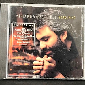 May include: A CD case featuring Andrea Bocelli's album "Sogno." The cover shows a close-up of Bocelli with his hands clasped. The album title and artist's name are at the top. A sticker indicates it's a "New Pop Album" with song titles and featured artists.