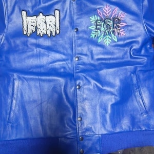 Könnte beinhalten: Blaue Lederjacke mit geripptem Kragen und Bündchen. Auf der linken Brust steht "FSRL", auf der rechten "FSR" mit Schneeflocken-Design. Die Jacke hat eine Knopfleiste und zwei Taschen.