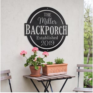 以下が含まれることがあります： THE Miller BACKPORCH Established 2019という白い文字が刻まれた円形の黒い金属製ウォールサイン。このサインは大胆でモダンなデザインとすっきりとしたフォントが特徴で、鉢植えが置かれた小さなテーブルの上の明るい色の壁に取り付けられています。