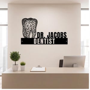 May include: A black metal wall sign featuring a geometric wireframe tooth design above the text DR. JACOBS and a rectangular bar below with the word DENTIST in white capital letters. The sign is mounted on a plain wall above a reception desk.