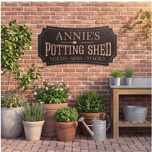 Puede incluir: Un letrero de metal negro con un borde decorativo que presenta el texto "ANNIE'S POTTING SHED" y "SEEDS • SOIL • TOOLS" en una fuente clara, con un icono de una pequeña pala entre las palabras Annie's y Potting.