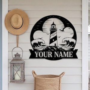 Pode incluir: Uma placa de parede circular em metal preto com um farol, ondas a quebrar e pássaros em silhueta. Abaixo do farol, uma faixa exibe o texto YOUR NAME em letras maiúsculas em negrito numa parede de cor clara.