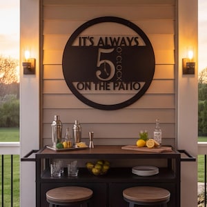 Puede incluir: Un letrero de pared circular de metal negro con el texto recortado IT'S ALWAYS 5 O'CLOCK ON THE PATIO. El letrero está montado en una pared de revestimiento horizontal beige sobre un carrito de bar marrón oscuro con cocteleras y fruta.