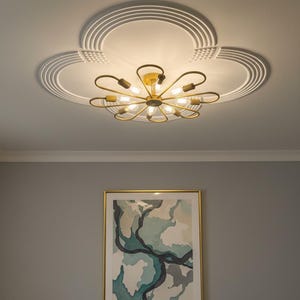 Puede incluir: Una lámpara de techo dorada con varias bombillas y un diseño floral. La luz está montada en un techo blanco con una moldura circular decorativa. Debajo, una pintura abstracta enmarcada cuelga de una pared gris.