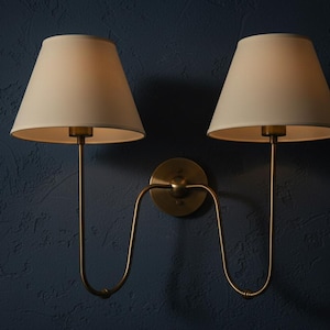 Puede incluir: Dos apliques de pared con pantallas de lámpara blanquecinas y accesorios metálicos de color latón. Los apliques tienen un diseño curvo y están montados en una pared azul oscuro texturizada. Las pantallas son cónicas.
