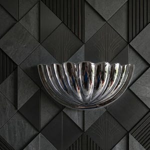 May include: A semi-circular wall sconce with a polished chrome finish and a scalloped, shell-like design. The fixture features deep vertical fluting that creates a reflective, curved surface, mounted against a dark, geometric tiled wall.