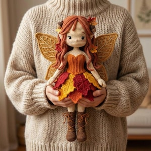 A handmade crochet autumn fairy doll with a light skin tone, wearing an orange corset and a layered red, yellow, and orange leaf skirt, with brown lace-up boots. The doll has long wavy auburn hair and four large golden leaf-shaped wings with embroidered vein details. The doll is being held up by a hand. A blurred outdoor autumn foliage background is behind.