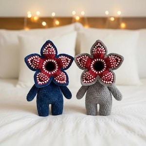 May include: Two handmade crochet Demogorgon plushies. One is navy blue, the other is gray. Both have a flower-like head with red and white detailing, and a black center. They are standing on a white surface.
