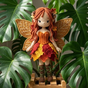 A handmade crochet autumn fairy doll with a light skin tone, wearing an orange corset and a layered red, yellow, and orange leaf skirt, with brown lace-up boots. The doll has long wavy auburn hair with acorn accessories and golden leaf-shaped wings. The doll is being cradled in two hands. A person wearing a cream knit sweater is holding the doll.