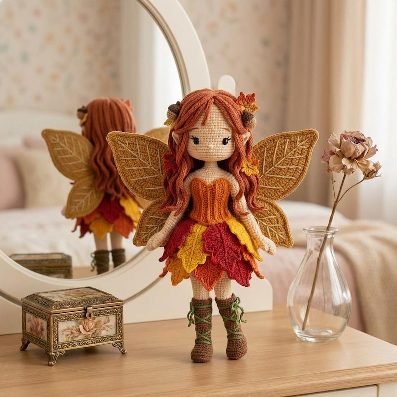 A handmade crochet autumn fairy doll with a light skin tone, wearing an orange corset and a layered red, yellow, and orange leaf skirt, with brown lace-up boots. The doll has long wavy auburn hair with acorn accessories and golden leaf-shaped wings. The doll is standing on a wooden vanity table. A round mirror and a glass vase with dried flowers are to the right.