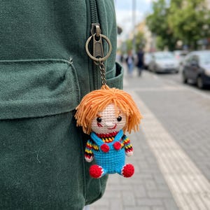 May include: A crochet doll keychain featuring a character with bright orange yarn hair, a pale face, and a red stitched smile. The doll wears blue overalls with red buttons over a multi-colored striped shirt and has red shoes, attached to a metal ring.
