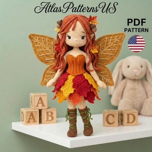May include: A crocheted fairy doll with red hair, autumn-colored leaf wings, and a dress made of red, yellow, and orange leaves. The doll is wearing brown boots and is displayed with wooden alphabet blocks and a stuffed bunny. The image includes the text "Atlas Patterns US" and "PDF PATTERN".