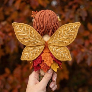 A handmade crochet autumn fairy doll with a light skin tone, wearing an orange corset and a layered red, yellow, and orange leaf skirt, with brown lace-up boots. The doll has long wavy auburn hair with acorn accessories and golden leaf-shaped wings. The doll is standing on a rustic wooden windowsill. Two terracotta herb pots are to the right.