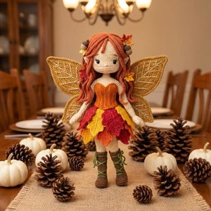 A handmade crochet autumn fairy doll with a light skin tone, wearing an orange corset and a layered red, yellow, and orange leaf skirt, with brown lace-up boots. The doll has long wavy auburn hair with acorn accessories and golden leaf-shaped wings. The doll is standing on a burlap table runner. White pumpkins and pine cones are surrounding the doll.