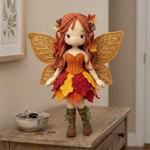 A handmade crochet autumn fairy doll with a light skin tone, wearing an orange corset and a layered red, yellow, and orange leaf skirt, with brown lace-up boots. The doll has long wavy auburn hair with acorn accessories and golden leaf-shaped wings. The doll is standing on a small wooden stand. Large monstera leaves are in the background.