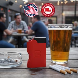 Puede incluir: Un encendedor rojo y negro con la letra "G" en el lateral está sobre una mesa de madera junto a un vaso de cerveza, cigarrillos y un cenicero. El fondo muestra a gente en una mesa al aire libre.