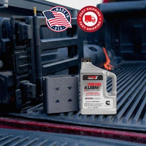 May include: A gray container and a bottle of Diesel Kleen fuel additive sit in the bed of a truck. The Diesel Kleen bottle is gray and white with black text and a black cap. The container is gray with a geometric design. The image includes a "Made in USA" sticker.