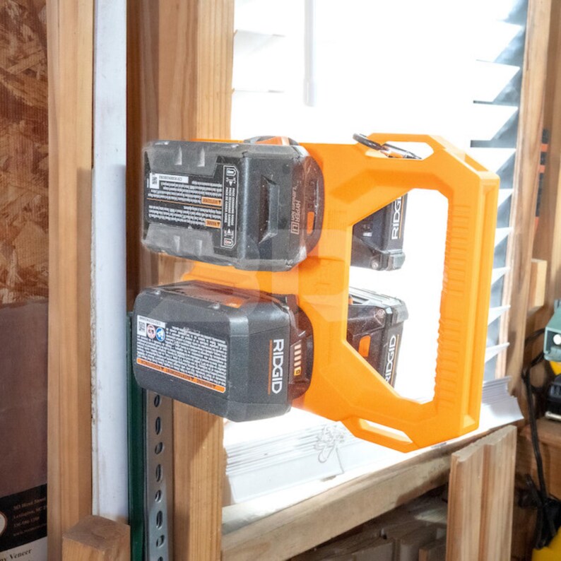 May include: An orange battery holder with two black Ridgid batteries attached. The holder is mounted on a wooden shelf near a window with white blinds. The holder has a handle for easy carrying.