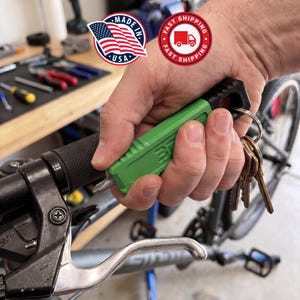 May include: A bright green multi-tool keychain featuring a black end cap and a metal screwdriver bit tip. The tool has a rectangular body with textured sides and a circular hole at the top for a metal keyring holding several silver keys.