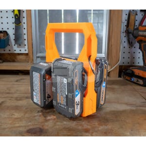 May include: An orange battery holder with a handle, holding three black and gray Ridgid 18V lithium-ion batteries. The batteries are labeled 4Ah and 3Ah. The holder is on a wooden surface.