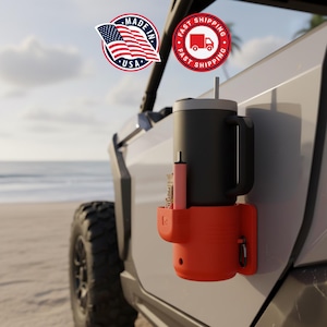 May include: A black insulated mug with a handle and a straw, mounted on an orange cup holder attached to a vehicle. A pink container and a carabiner are also visible. The image includes a "Made in USA" sticker and a "Fast Shipping" sticker.