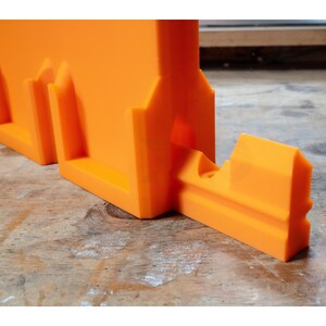 May include: Bright orange plastic tool with a rectangular base and angled supports. The tool has a slot for holding a piece of material and is designed for cutting or shaping. The tool is sitting on a wooden surface.