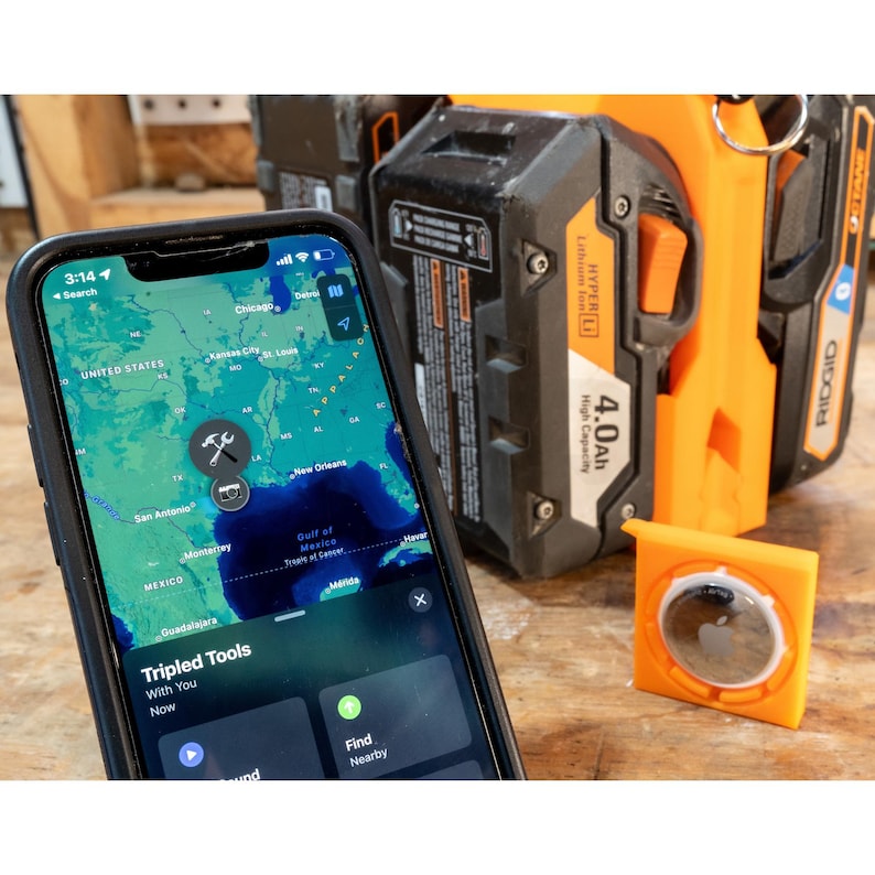 May include: A smartphone displaying a map with the text "Tripled Tools" and a wrench icon. An orange AirTag holder and a black and orange power tool battery are in the background.