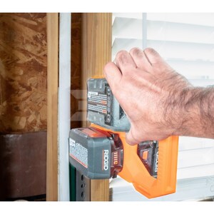 May include: An orange and black RIDGID power tool with two attached batteries. The tool is being held by a person's hand near a wooden wall and a window with white blinds. The batteries have warning labels.