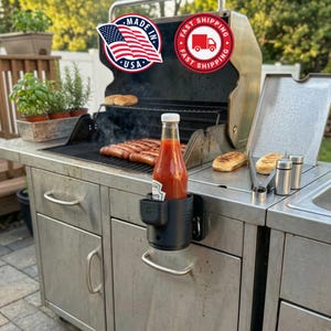 May include: A black cup holder with a bottle of ketchup attached to a stainless steel outdoor grill. The grill has sausages cooking on it. The image includes a "Made in USA" sticker and a "Fast Shipping" sticker.