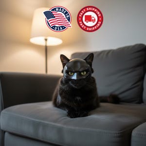 Puede incluir: Un gato negro con una máscara negra y una expresión severa, sentado en un sofá gris. El gato tiene ojos verdes. La imagen incluye una pegatina "Made in USA" y una pegatina "Fast Shipping".