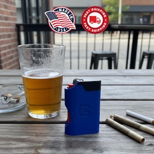 May include: A blue and red lighter with a black top and the letter "G" on the side sits on a wooden table next to a glass of beer, an ashtray, and rolled cigarettes. The image includes a "Made in USA" sticker.