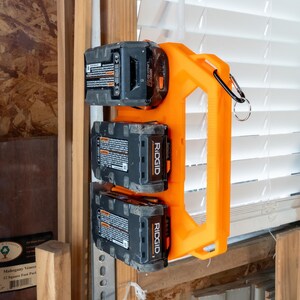 May include: An orange power tool battery holder with three black and gray Ridgid batteries attached. The holder is mounted on a wooden structure near a window with white blinds. A black carabiner is attached to the holder.