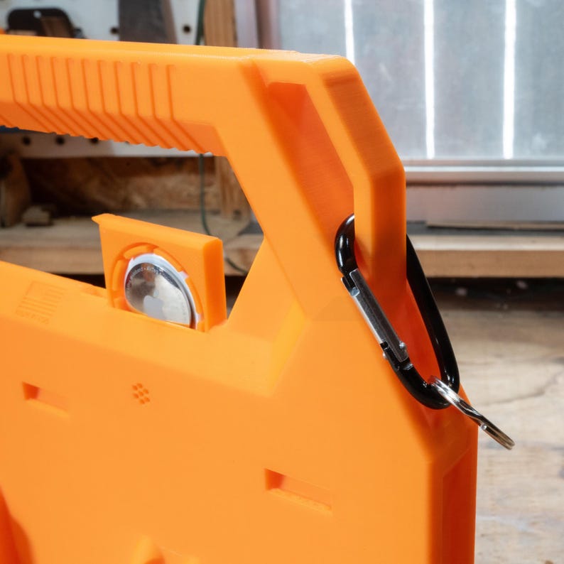 May include: Close-up of an orange plastic tool with a handle and a black carabiner clip attached. The tool has a rectangular shape with a small, circular, reflective element and several rectangular cutouts.