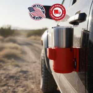 May include: A red cup holder attached to a black vehicle door, holding a stainless steel tumbler. The cup holder has a small side compartment. The background shows a dirt road and desert landscape. The image includes "Made in USA" and "Fast Shipping" logos.