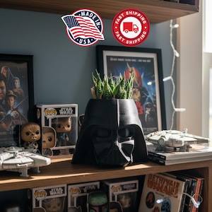 May include: A black Darth Vader helmet-shaped planter filled with green succulents. The planter sits on a wooden shelf with Star Wars figurines, movie posters, and a model of the Millennium Falcon. The image also includes a "Made in USA" sticker.