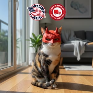 May include: A calico cat wearing a red devil mask with horns, sitting on a wooden floor. The cat has a white chest and face, with black, brown, and orange markings on its fur. The background includes a window, a plant, and a couch.
