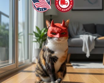 May include: A calico cat wearing a red devil mask with horns, sitting on a wooden floor. The cat has a white chest and face, with black, brown, and orange markings on its fur. The background includes a window, a plant, and a couch.