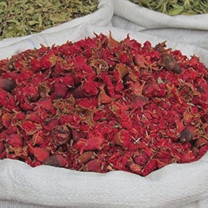 May include: A white sack overflowing with dried red pomegranate flowers. The flowers are a deep red colour with brown and tan highlights. The image suggests a natural product, possibly for culinary or medicinal purposes.