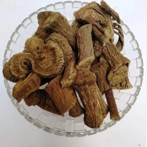 Può includere: Una ciotola di vetro trasparente piena di pezzi marroni secchi di forma irregolare, forse una radice o un'erba. I pezzi variano per dimensioni e consistenza, alcuni mostrano un motivo circolare a strati. La ciotola è su una superficie bianca.