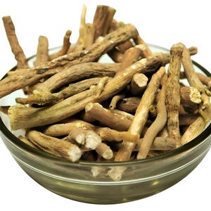 Op de afbeelding: Een heldere glazen kom gevuld met gedroogde, lichtbruine ashwagandha-wortels. De wortels zijn van verschillende grootte en vorm, met een natuurlijke, aardse uitstraling. De kom staat tegen een witte achtergrond.